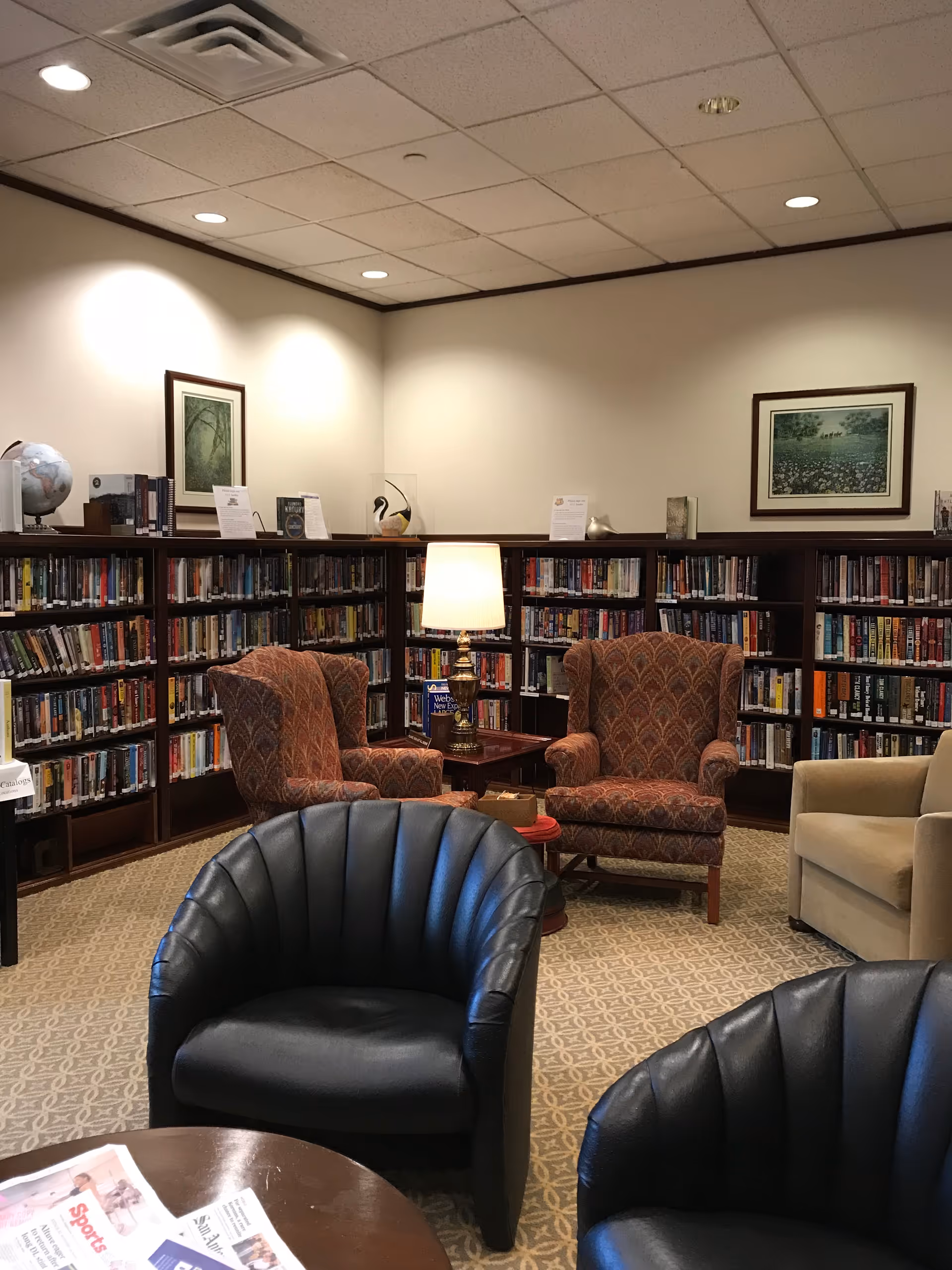 Cozy library room with bookshelves lining the walls filled with books. There are several comfortable chairs including two patterned armchairs, a beige armchair, and two black leather chairs around a wooden table with newspapers. A table lamp is placed on a small table between the patterned armchairs. The walls have framed artwork and the ceiling has recessed lighting.