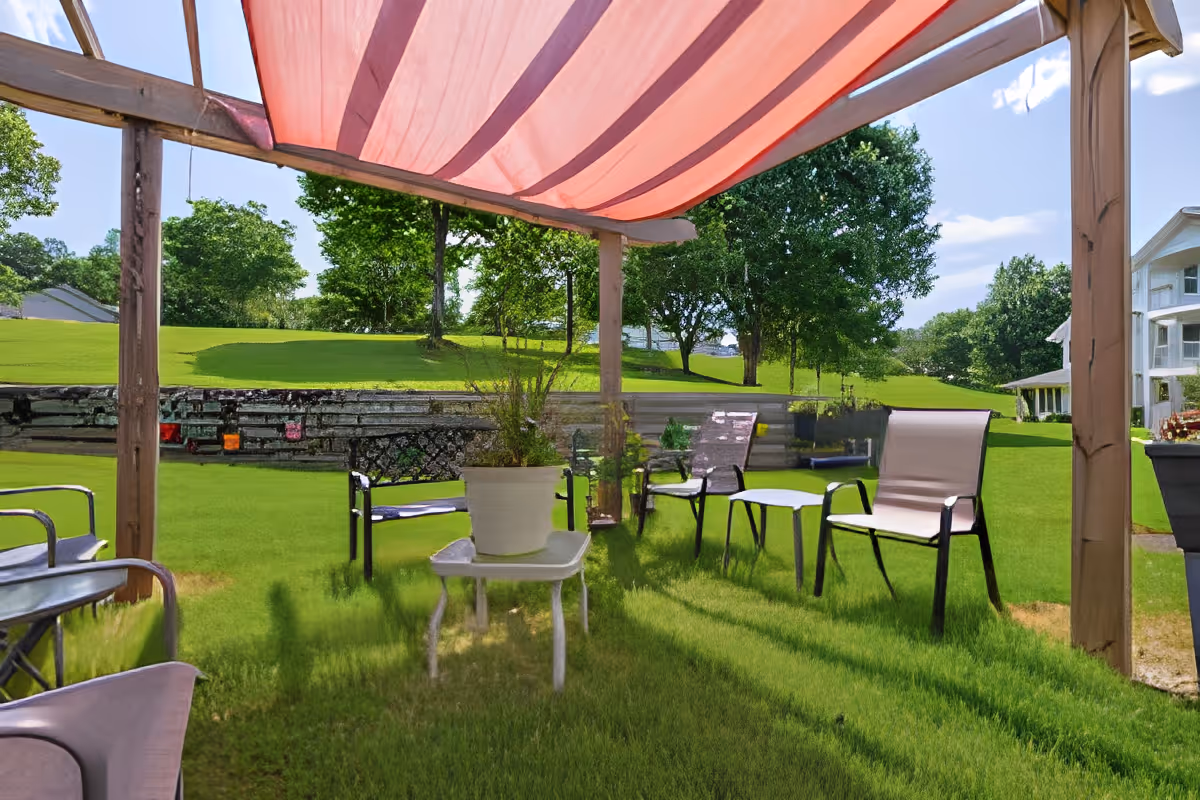 Patio seating area with chairs and a small table holding a potted plant under a pink canopy overlooking a grassy lawn and trees.