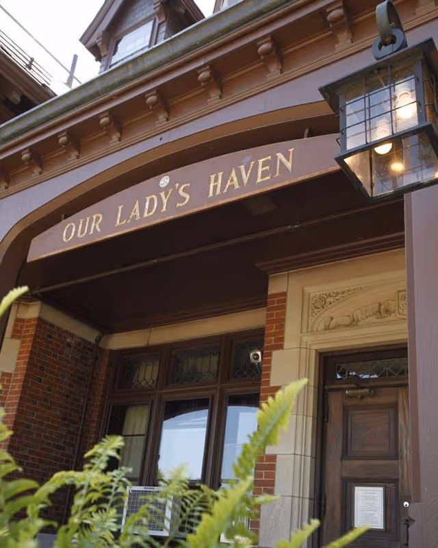 Entrance of a building with a sign above the door that reads 'OUR LADY'S HAVEN'. The entrance features a wooden door with decorative stonework around it, brick walls, and a lantern-style light fixture on the right side. Green plants are visible in the foreground.