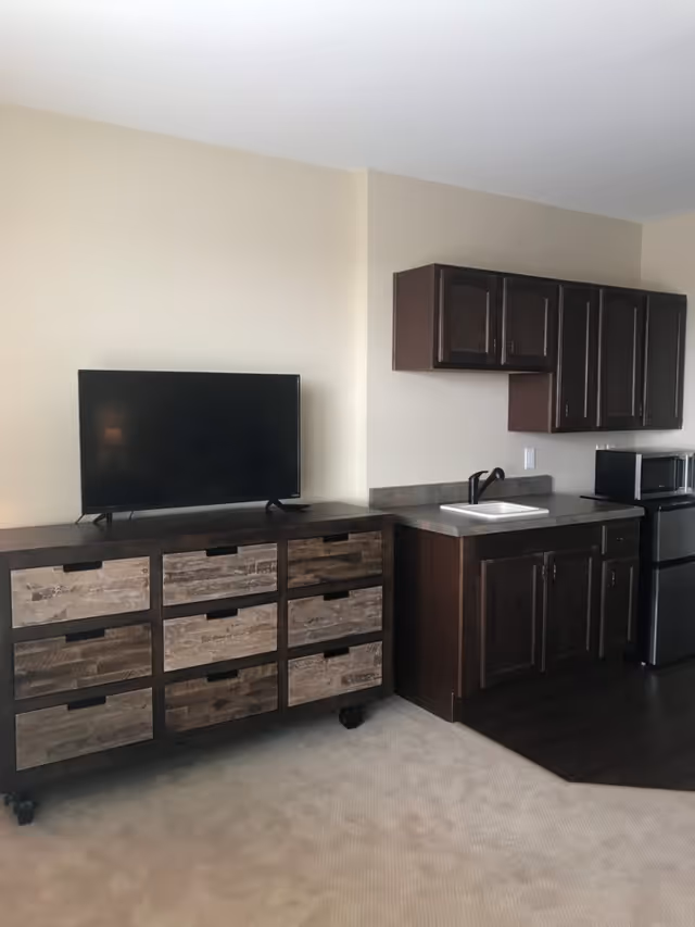 Interior view of a room featuring a wooden dresser with multiple drawers on the left, a flat-screen TV on top of the dresser, and a kitchenette area on the right with dark wood cabinets, a countertop with a sink, a microwave, and a mini refrigerator.