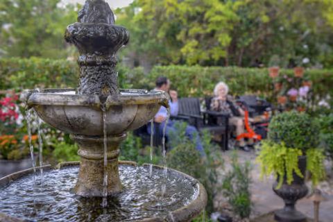 A stone water fountain with water flowing from the top tier to the lower basin in a garden setting. In the background, two elderly people are seated on benches surrounded by lush greenery and colorful flowers.