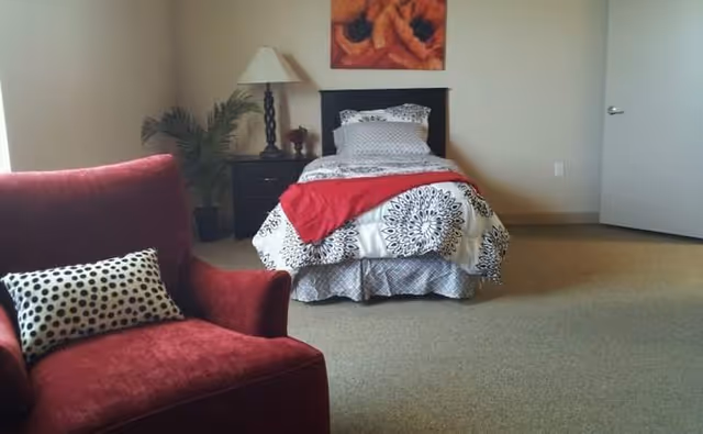 A bedroom with a patterned bed, red throw, nightstand, lamp, and red chair.