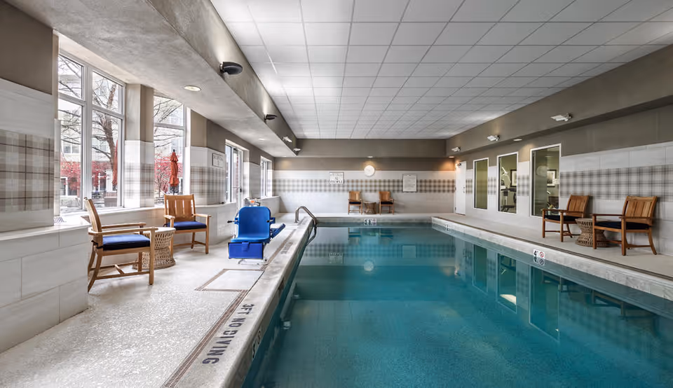 Indoor swimming pool area with seating, a pool lift, large windows and tiled walls.