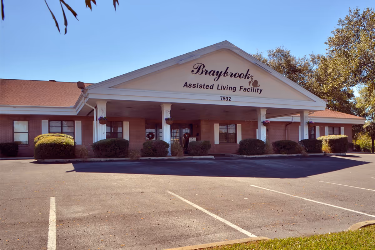 Exterior front view of Braybrook Assisted Living Facility, a single-story brick building with a covered entrance supported by white columns, surrounded by bushes and trees under a clear blue sky.