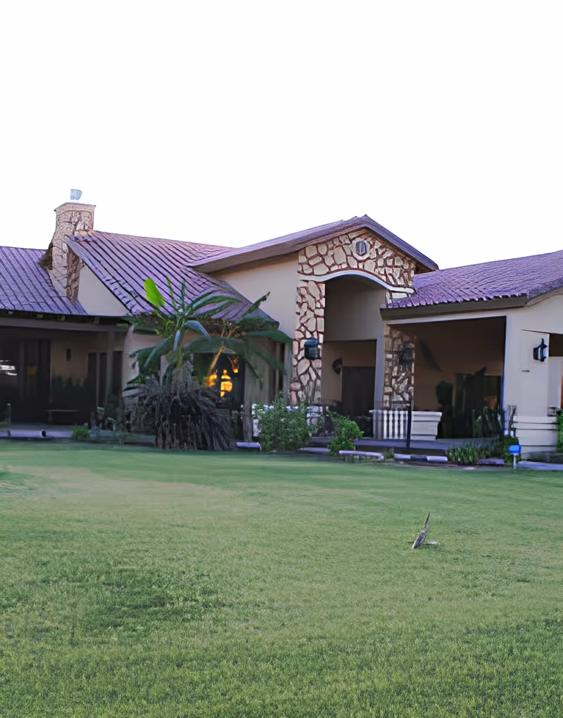 Exterior view of a single-story building with a stone and stucco facade, a red-tiled roof, and a well-maintained green lawn in front. There are plants and shrubs near the building, and the entrance features an archway with stone detailing.