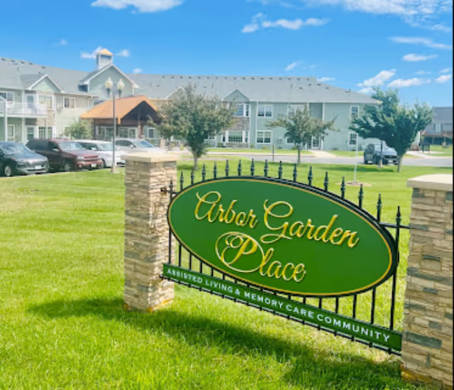 A green and gold sign reading 'Arbor Garden Place Assisted Living & Memory Care Community' is mounted on a black iron fence supported by two stone pillars, with a large building, parked cars, trees, and a bright blue sky in the background.