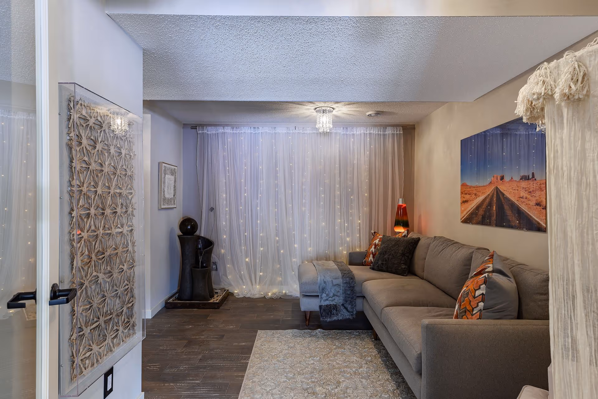 A cozy living room with a gray sectional sofa adorned with patterned pillows and a gray throw blanket. A lava lamp sits on a side table next to the sofa. Sheer white curtains with string lights cover the window behind the sofa. A large wall art piece depicting a desert road scene hangs above the sofa. The floor is dark wood, and a light-colored patterned rug is partially visible. Decorative elements include a sculptural piece in the corner and textured wall art near the door.