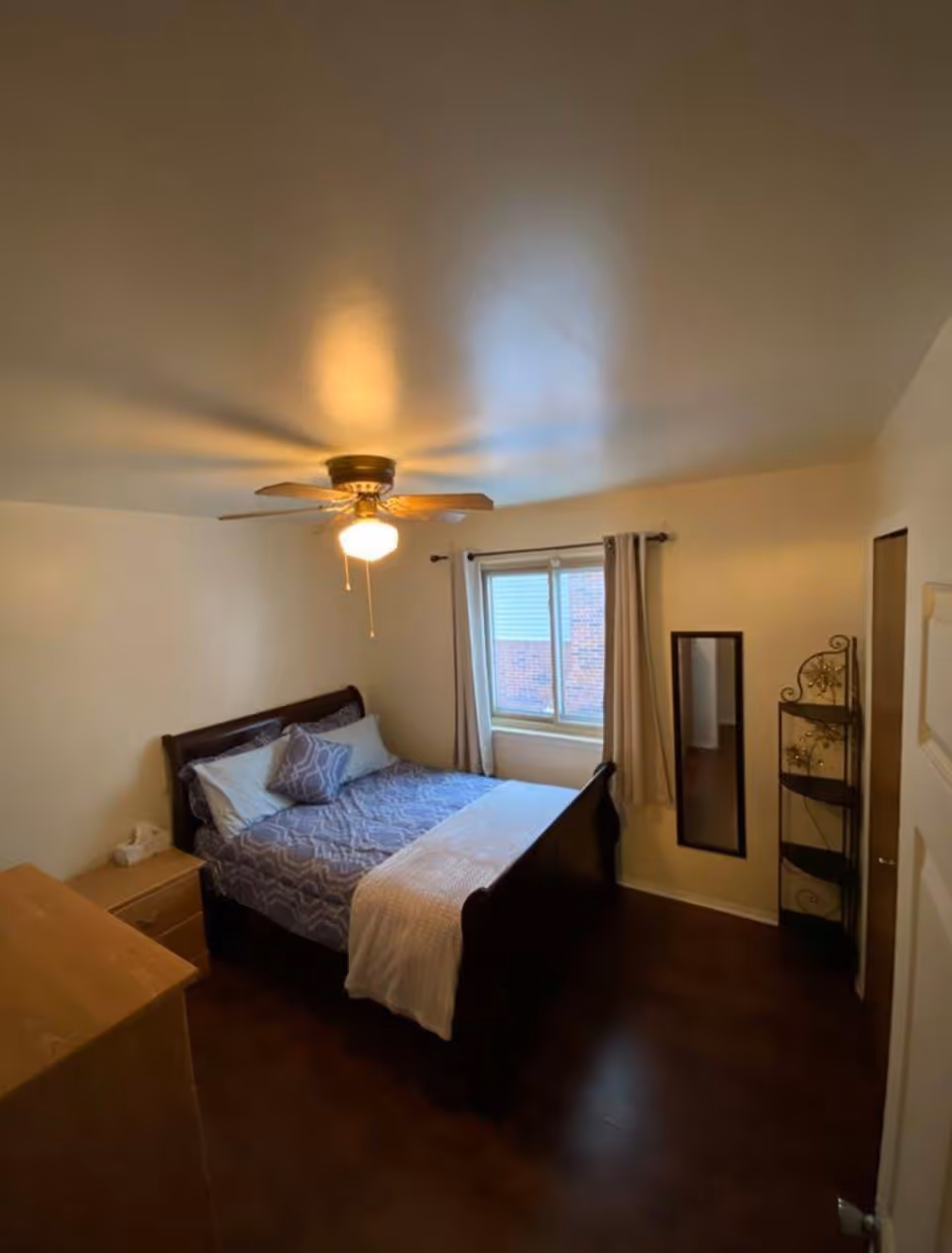 Bedroom with a dark wood bed, ceiling fan, window with curtains, dresser and corner shelving.