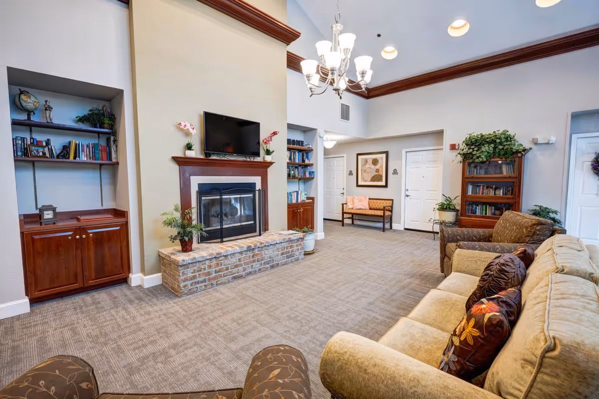 A cozy living room area in a senior living facility with a beige sofa adorned with floral pillows, a brown armchair, a brick fireplace with a mounted flat-screen TV above it, and built-in wooden bookshelves on either side. The room has a chandelier, carpeted floor, and additional seating with a bench near the back wall, decorated with plants and framed artwork.