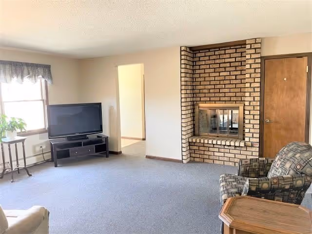 A living room with a brick fireplace, a flat-screen TV on a black TV stand, a window with a valance, a patterned armchair, a wooden side table, and a beige armchair partially visible.