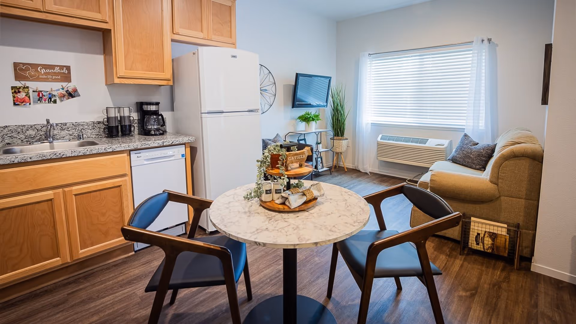 Open studio apartment with a kitchenette, round marble dining table and chairs, and a living area with a sofa and wall-mounted TV.