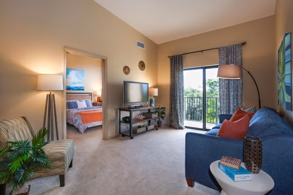 A cozy living room with a blue sofa adorned with red pillows, a round side table with books and a decorative vase, a floor lamp, and a TV on a stand. The room has beige walls and carpet, with a sliding glass door leading to a balcony with greenery outside. Through an open doorway, a bedroom with a bed, bedside lamps, and a blue and orange bedspread is visible.