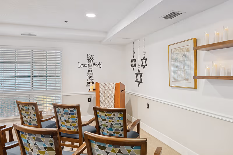 A small room with several wooden chairs featuring geometric patterned cushions arranged facing a wooden podium draped with a cloth. The walls are decorated with hanging Star of David symbols, a framed picture, and a wall decal with a religious quote. A window with blinds allows natural light into the room, and shelves on the wall hold multiple candles.