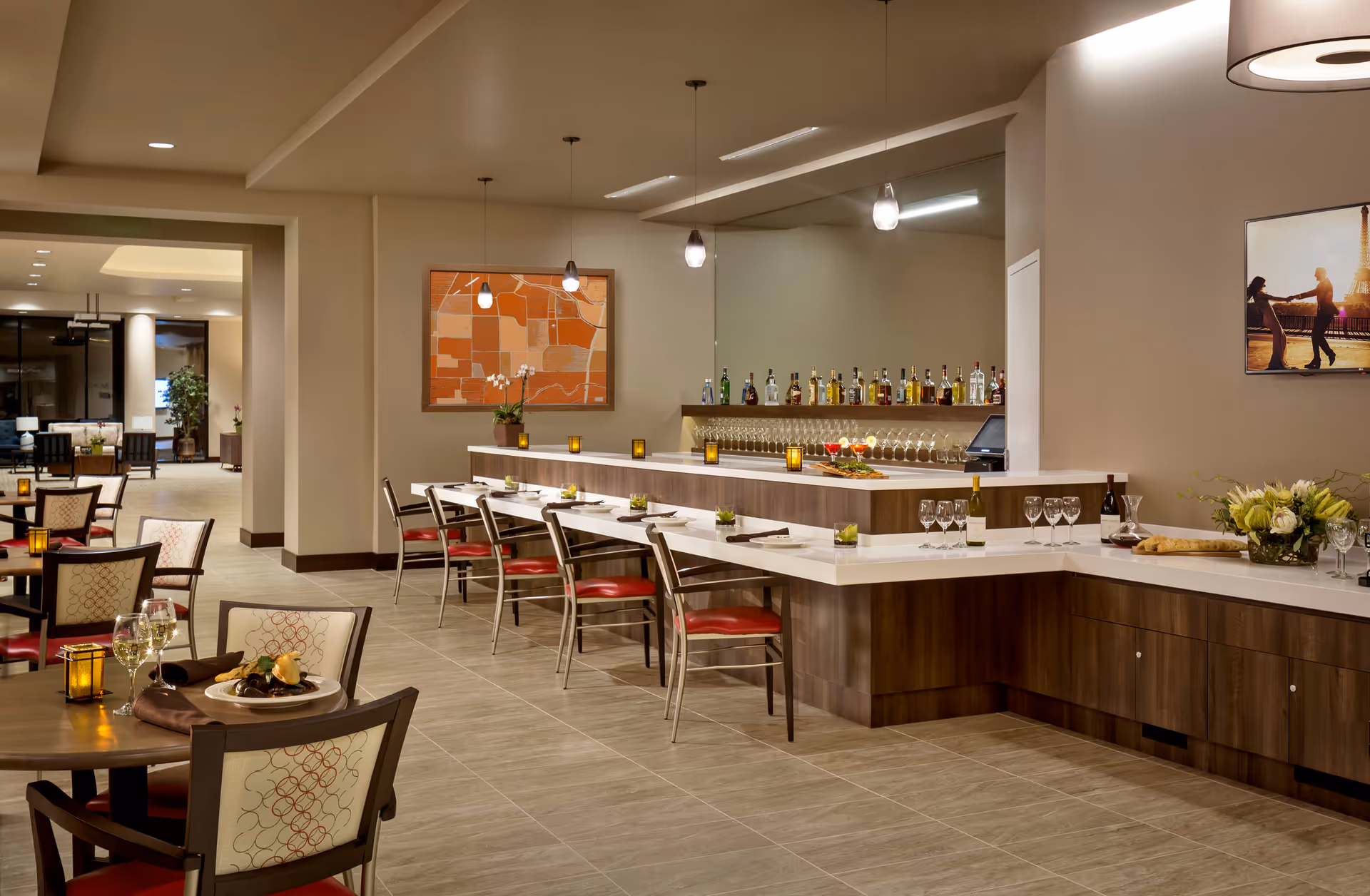 A modern dining area in a senior living facility featuring a long bar with bottles and glasses, several tables with chairs, and warm lighting. The space has tiled floors, contemporary decor, and a cozy atmosphere.