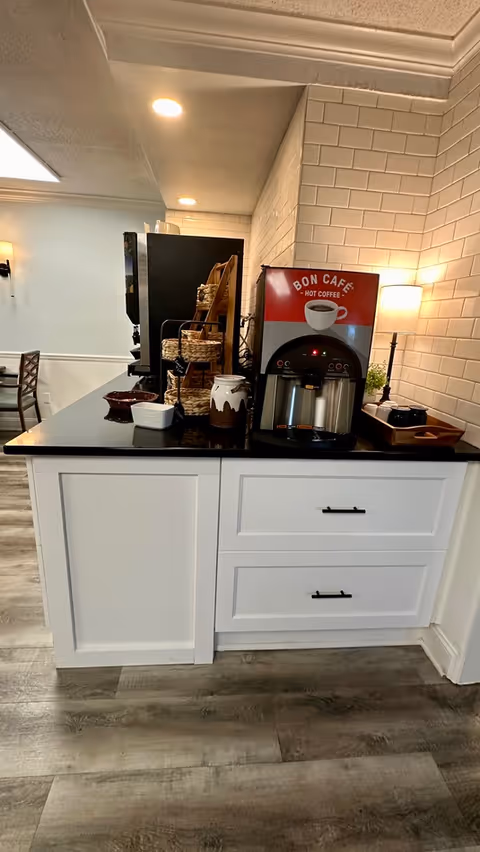 A coffee station in a room with white tiled walls and wood flooring. The station features a Bon Cafe hot coffee machine on a black countertop with white cabinetry below. Next to the coffee machine are baskets, a jar, and a small lamp on a wooden tray. In the background, part of a dining area with chairs and tables is visible.