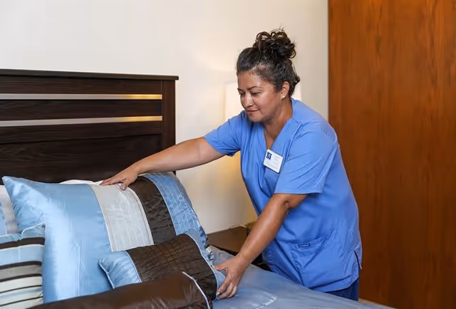 A caregiver in blue scrubs is making a bed with blue and brown pillows in a bedroom setting.