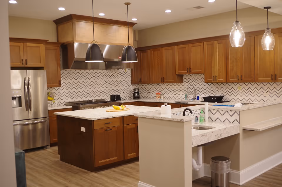 Modern kitchen with wooden cabinets, a stainless steel refrigerator, a central island with a fruit bowl, pendant lights hanging from the ceiling, and a white and gray chevron-patterned backsplash.