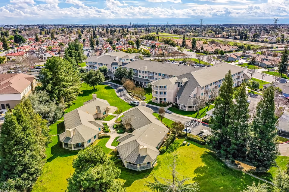 Aerial view of Solstice Senior Living at Bakersfield showing multiple residential buildings surrounded by green lawns, trees, and pathways, with a suburban neighborhood in the background under a partly cloudy sky.