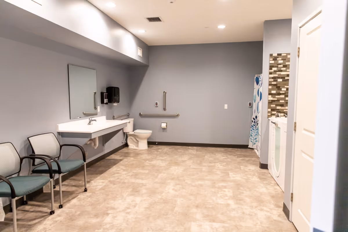 A spacious bathroom with light gray walls and beige tiled floor. The room features a wall-mounted sink with a mirror above it, a toilet with grab bars on the wall, two chairs with armrests, and a shower area with a blue and white patterned curtain. There is also a washing machine and dryer next to the shower area.