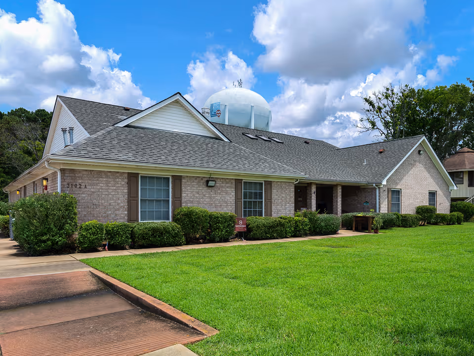 Exterior view of a single-story brick building with a gray shingled roof, surrounded by green grass and bushes under a partly cloudy blue sky. A water tower is visible in the background.