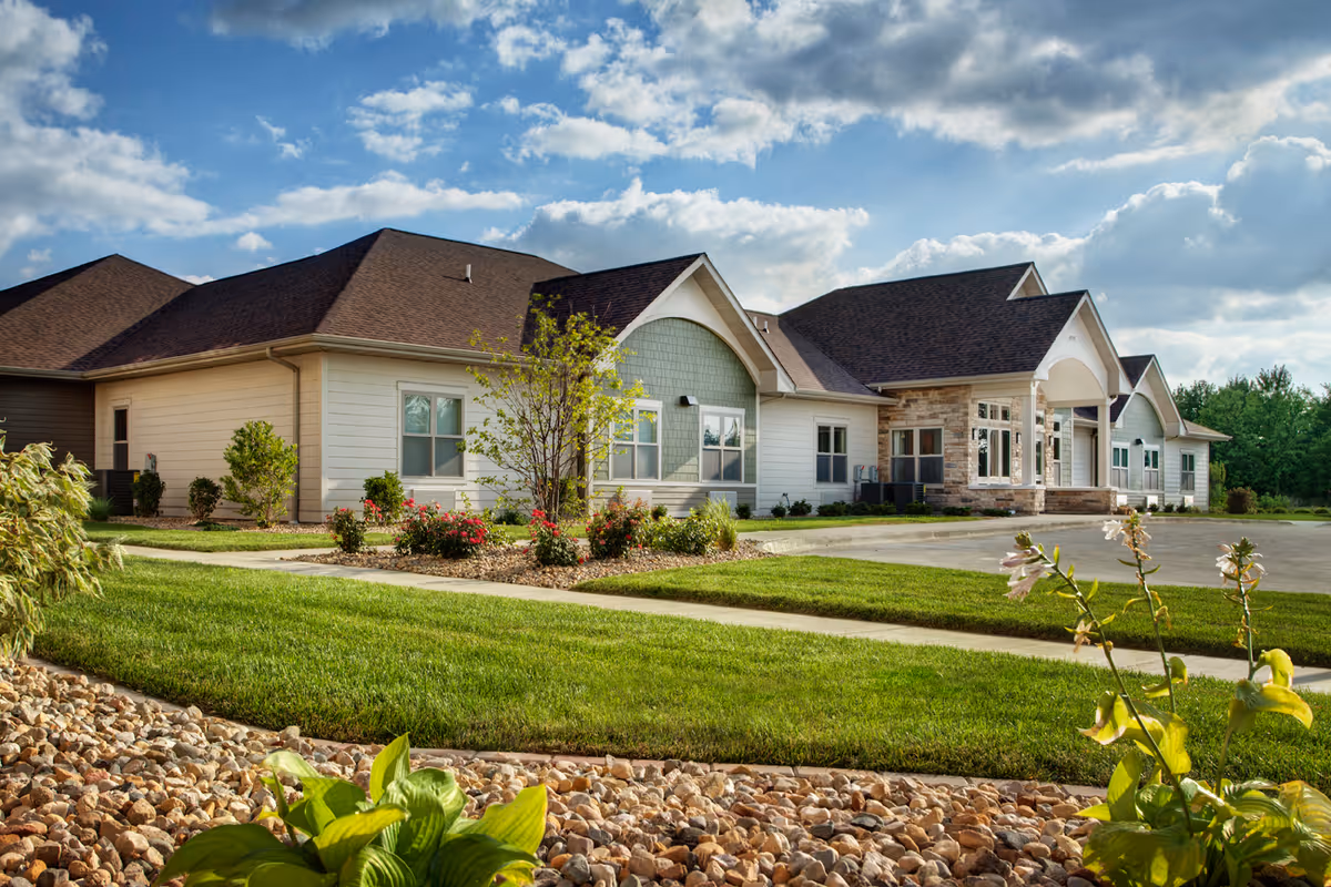 Exterior view of a single-story senior living facility building with a well-maintained lawn, landscaped garden beds with flowers and shrubs, a paved driveway, and a partly cloudy blue sky.