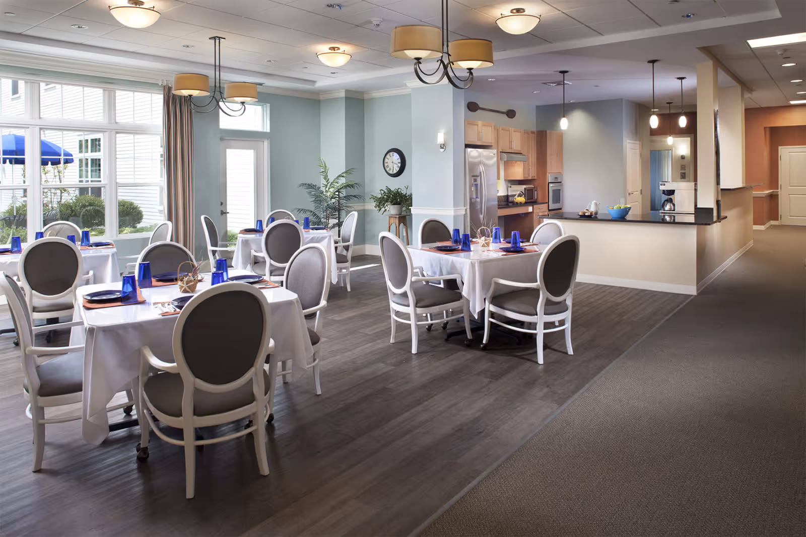 A bright dining room with several round tables covered with white tablecloths, each set with blue glasses and napkins. The room features gray cushioned chairs, large windows letting in natural light, and a view of an outdoor patio with blue umbrellas. In the background, there is a kitchen area with wooden cabinets, stainless steel appliances, and a countertop with a bowl of fruit.