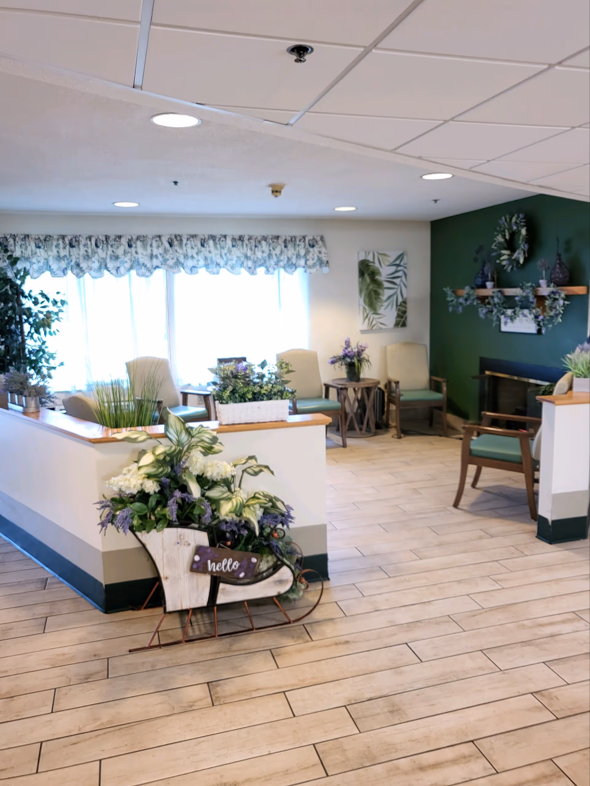 A bright and welcoming common area with light wood flooring, several chairs arranged around small tables, and a decorative green wall with shelves and a wreath. A large window with floral curtains lets in natural light. In the foreground, there is a decorative planter shaped like a sled filled with white and purple flowers and a small sign that says 'hello'.