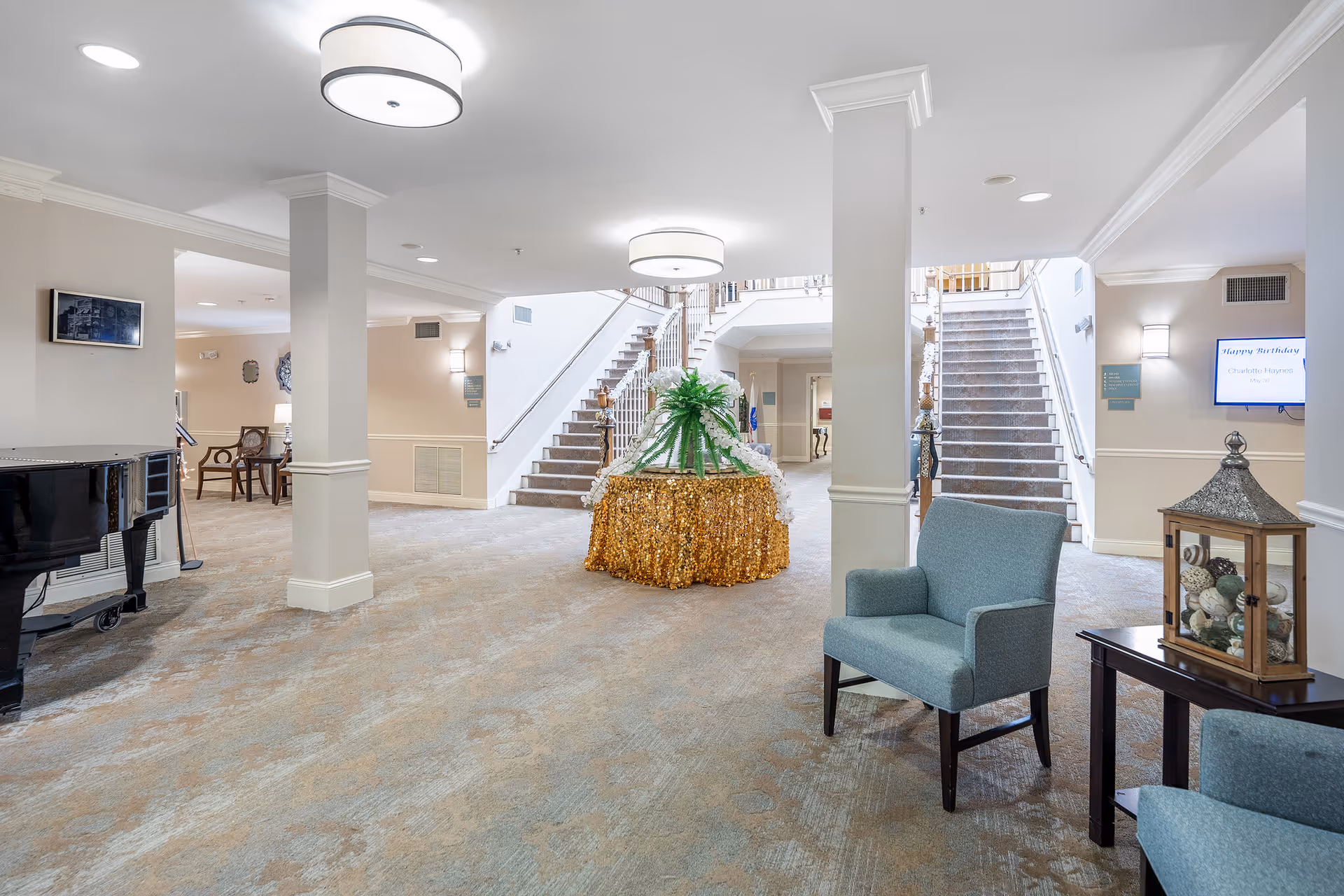 Interior view of a senior living facility lobby with carpeted floors, two staircases leading to an upper level, a round table with a gold sequin tablecloth and a green plant centerpiece, blue upholstered chairs, a black grand piano on the left, and wall-mounted lights and screens.