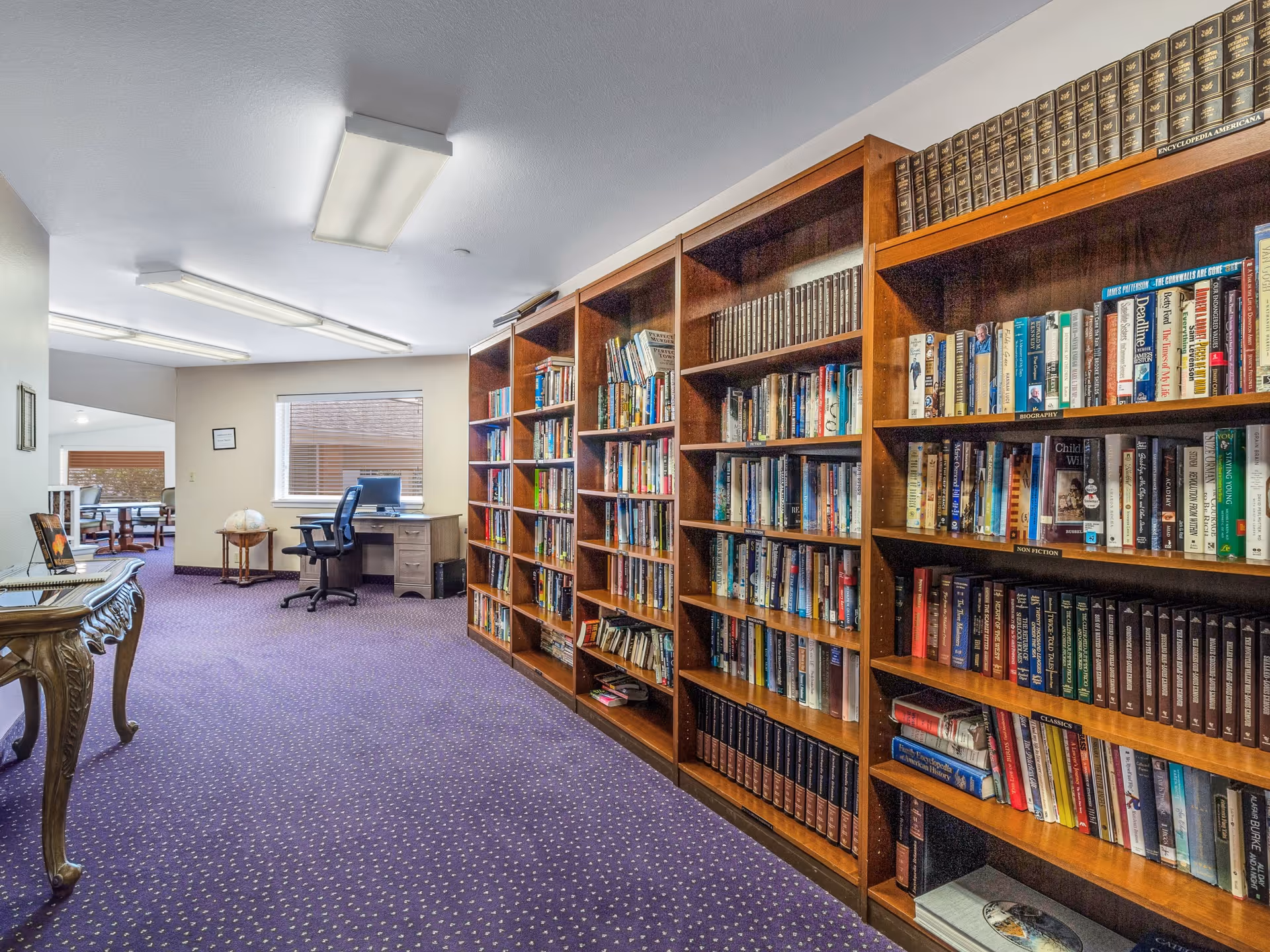 Interior view of a senior living facility library area with wooden bookshelves filled with books on the right side, a purple carpet with small patterns, a wooden table with decorative legs on the left, and a desk with a computer and office chair near a window in the background.