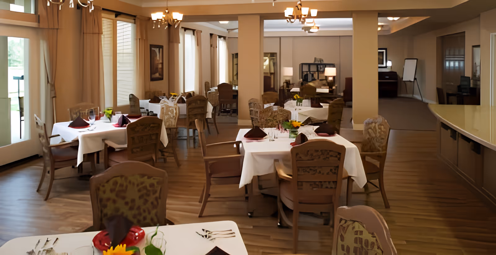 Bright, upscale dining room with round tables dressed in white linens, folded napkins, cushioned wooden chairs, chandeliers, and wood flooring.