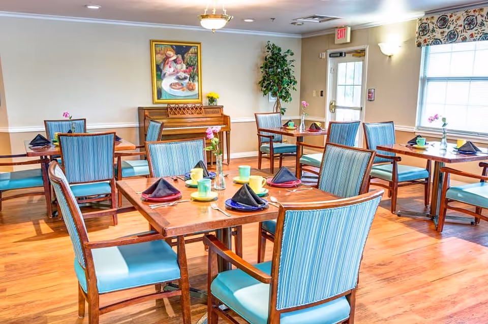 A bright dining room with wooden tables and chairs featuring blue striped upholstery. Each table is set with colorful cups, saucers, and black napkins folded in a triangular shape. A piano with a framed painting above it and a tall potted plant are visible against the far wall. Large windows allow natural light to fill the room.