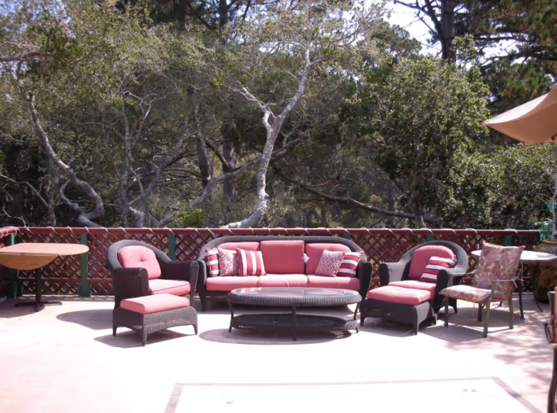 Outdoor patio area with cushioned wicker furniture including a sofa, two armchairs, and a coffee table. There are also two small round tables with chairs. The patio is surrounded by a wooden lattice fence and trees in the background.