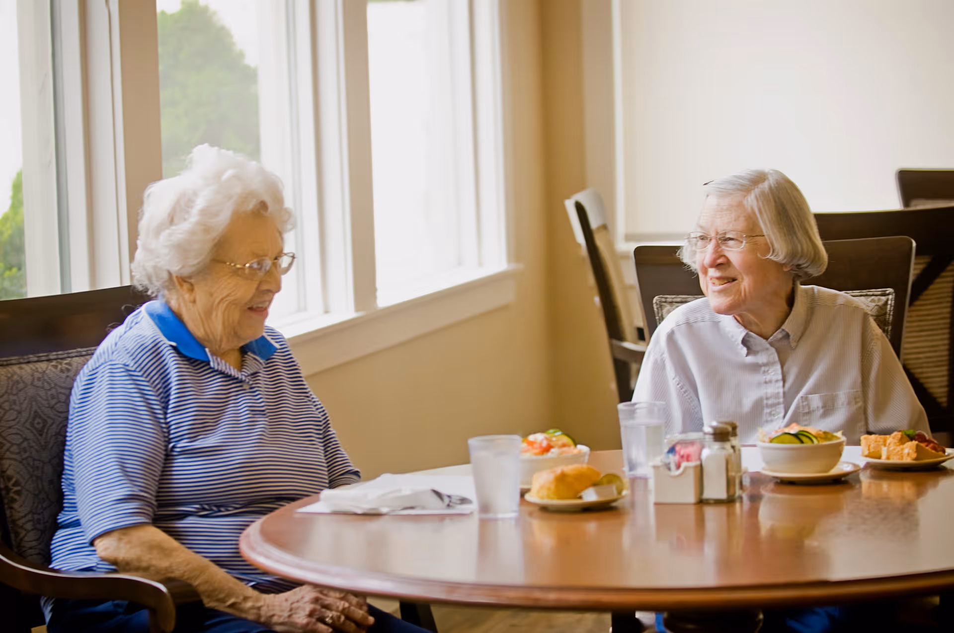 Two elderly women sitting at a round wooden dining table in a well-lit room, smiling and engaging in conversation. The table has plates with food, glasses of water, and condiments. Large windows with white trim allow natural light to fill the space.