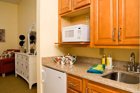 Small kitchenette with a microwave, mini-refrigerator, sink and granite countertop, with a dresser and seating visible in an adjacent living area.