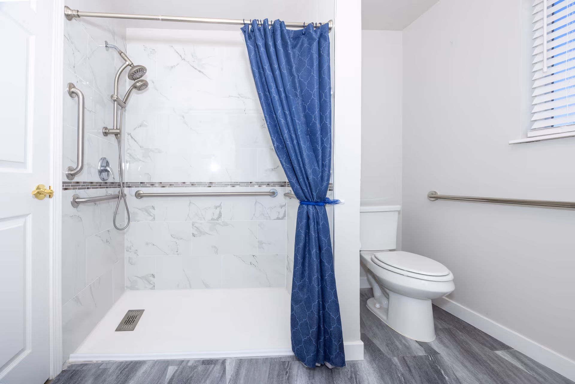A clean, modern bathroom featuring a walk-in shower with white marble-patterned tiles, a blue shower curtain tied to one side, stainless steel grab bars, and a handheld showerhead. To the right, there is a white toilet next to a wall with a horizontal grab bar and a window with white blinds. The floor has gray wood-like tiles.