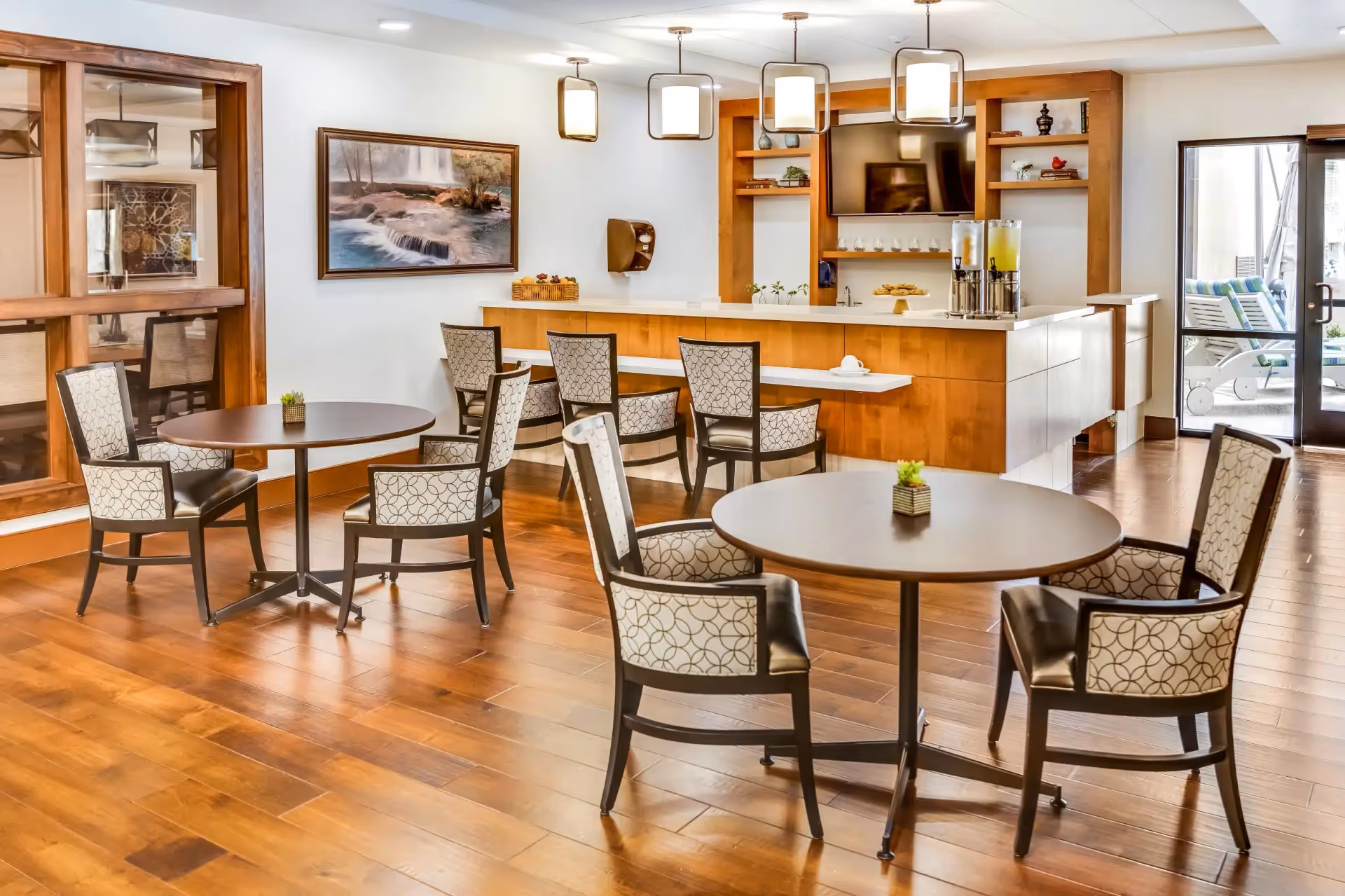 Bright communal dining area with round tables and upholstered chairs facing a wooden service counter with beverage dispensers and a TV.