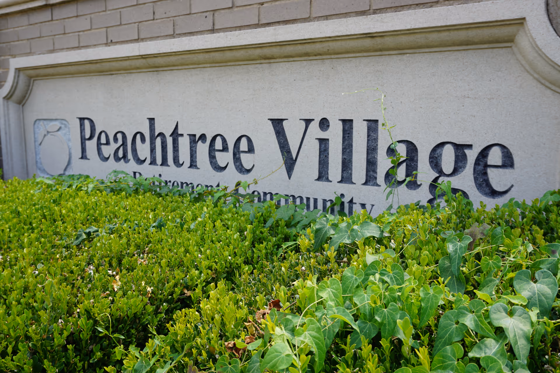 Stone sign for Peachtree Village Retirement Community partially covered by green bushes and ivy, mounted on a brick wall.