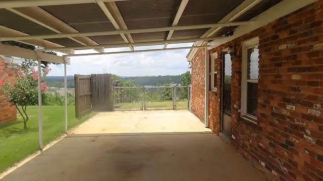 Covered patio/carport attached to a brick house overlooking a fenced backyard and distant trees.