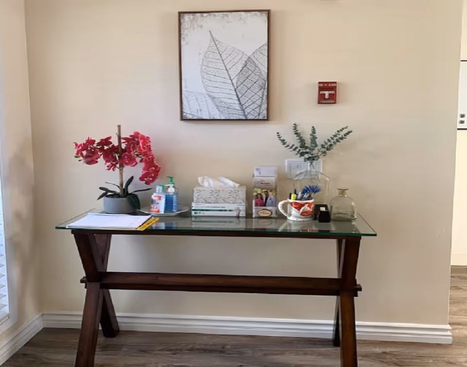 A wooden table with a glass top against a beige wall, decorated with a potted pink orchid, hand sanitizer, tissue box, brochures, a mug with pens, a small glass bottle, and a vase with green foliage. Above the table is a framed artwork of leaf outlines and a red fire alarm on the wall.