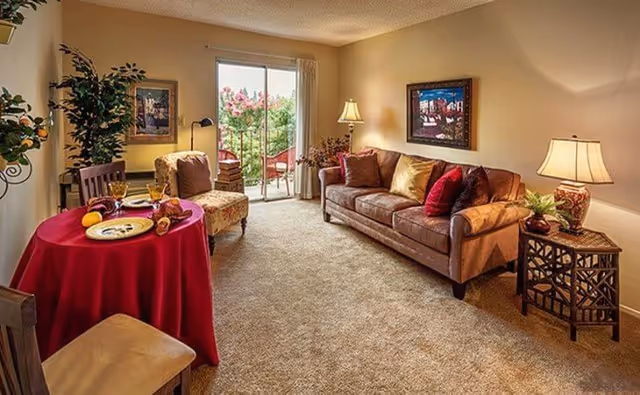 Cozy living room with a sofa, armchair, small round table with a red tablecloth, side tables and a sliding glass door to a balcony.