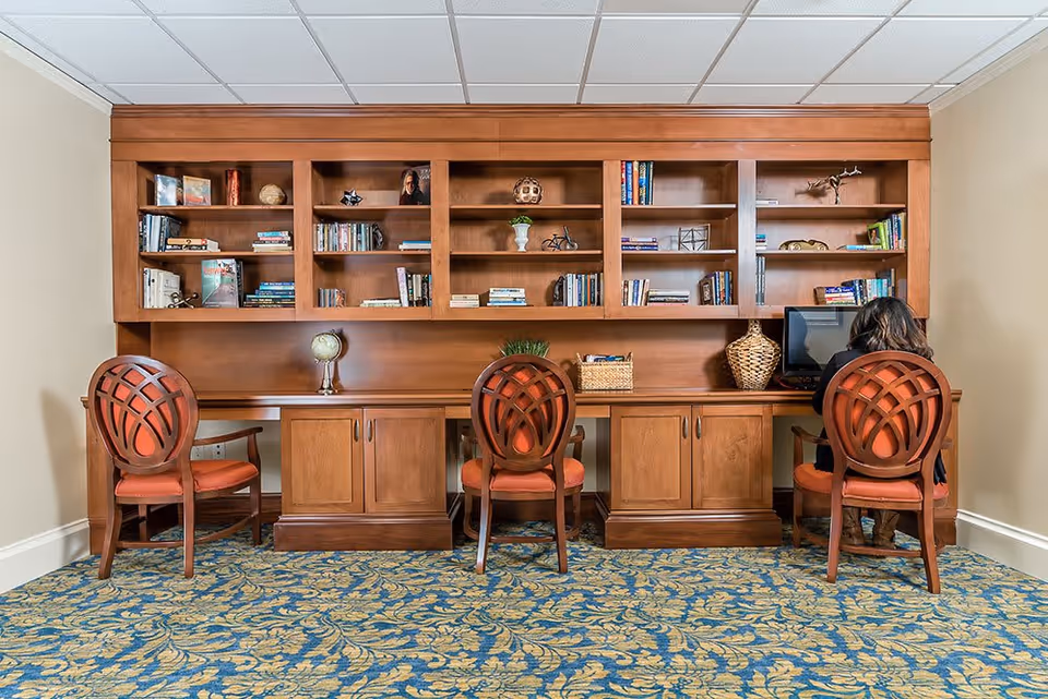 A study or computer area with three wooden chairs with orange cushions placed in front of a long wooden desk. The desk has built-in cabinets and shelves above it filled with books and decorative items. A person is seated on the right chair using a computer. The room has a patterned blue and gold carpet and beige walls.