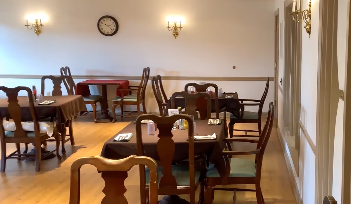 Empty dining room with wooden tables and chairs set with brown tablecloths, place settings, wall sconces, and a clock on the wall.