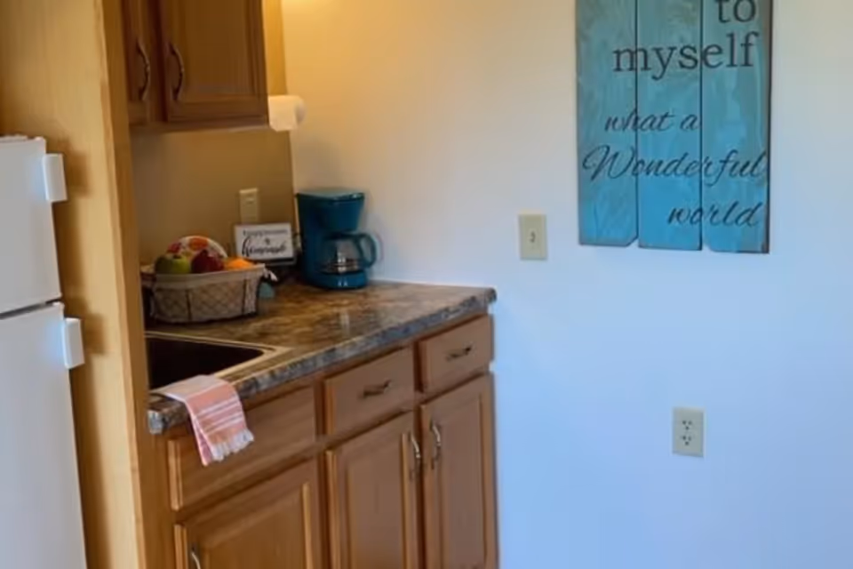 A small kitchen area with wooden cabinets, a countertop with a coffee maker, a basket of fruit, and a sink. A white refrigerator is partially visible on the left. On the wall, there is a decorative sign with the words 'to myself what a Wonderful world'.