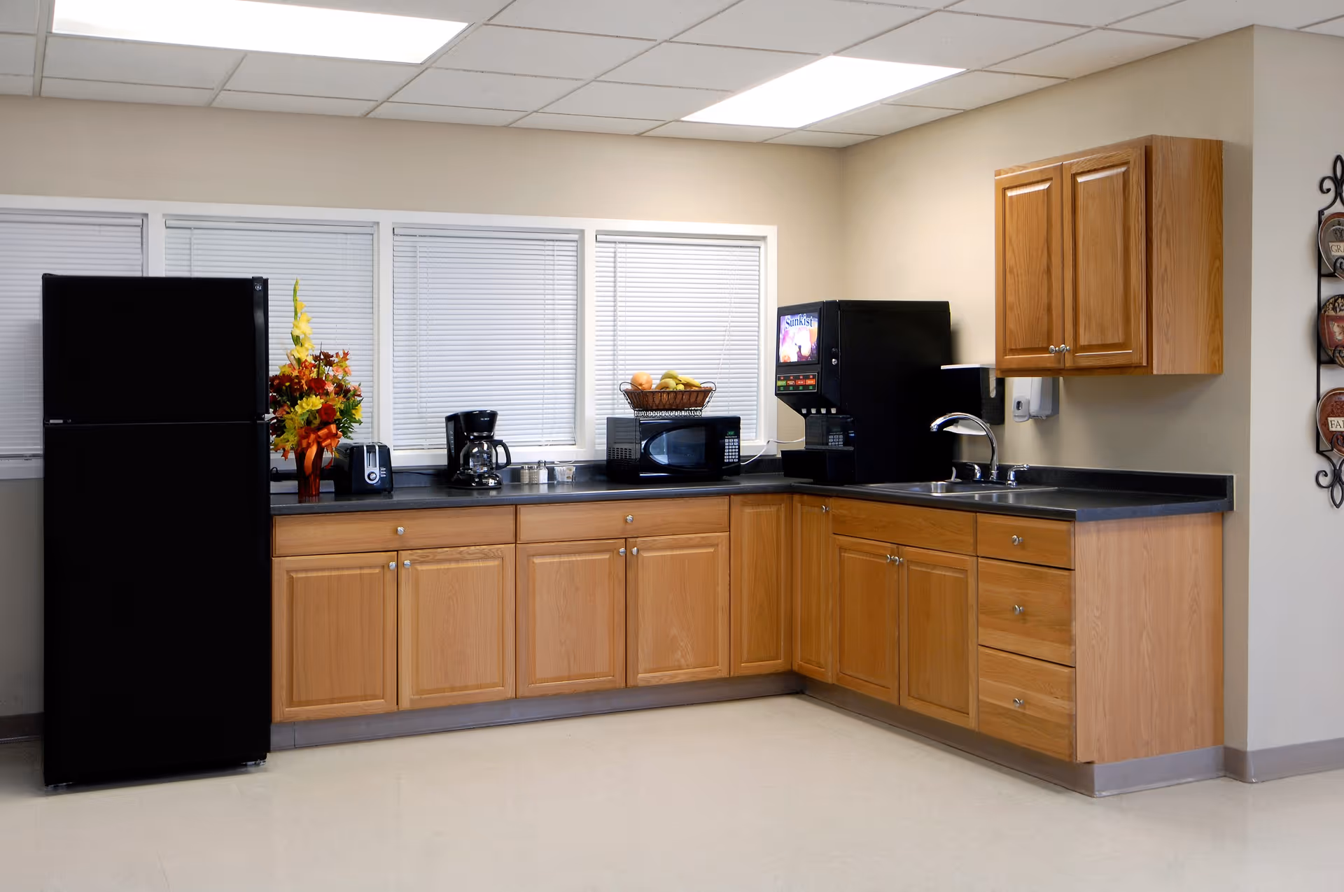 Communal kitchenette with wooden cabinets, a black refrigerator, countertop appliances (microwave, coffee maker, beverage dispenser), a sink and a bowl of fruit.