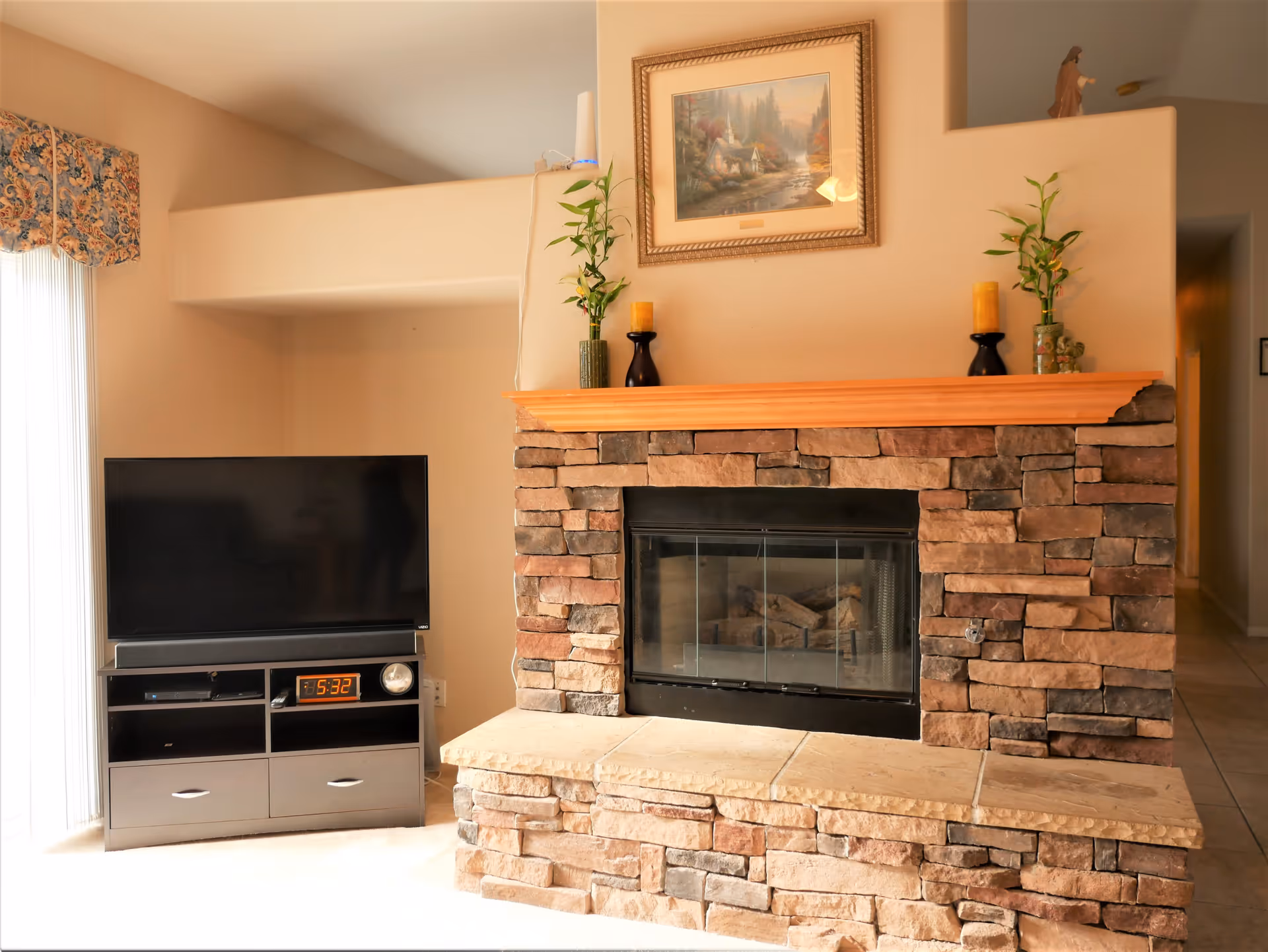 A cozy living room area featuring a stone fireplace with a wooden mantel decorated with two candles and two small plants. Above the mantel hangs a framed painting of a cabin in the woods. To the left of the fireplace is a flat-screen TV on a stand with shelves and drawers, displaying a digital clock showing 5:32. The room has beige walls and a window with floral valance curtains letting in natural light.