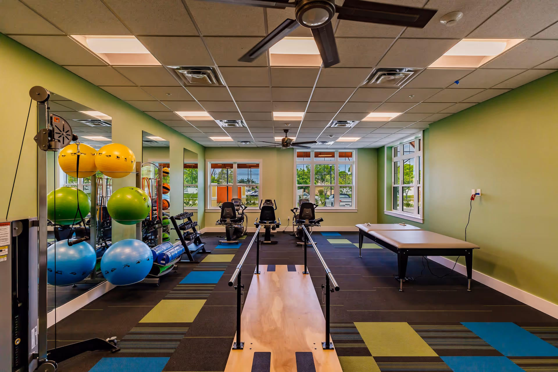 A well-lit fitness room with green walls and large windows. The room contains exercise equipment including stability balls in yellow, green, and blue, dumbbells on racks, two recumbent exercise bikes, a padded therapy table, and parallel bars for walking exercises. The floor has a patterned carpet with blue, green, and black squares.