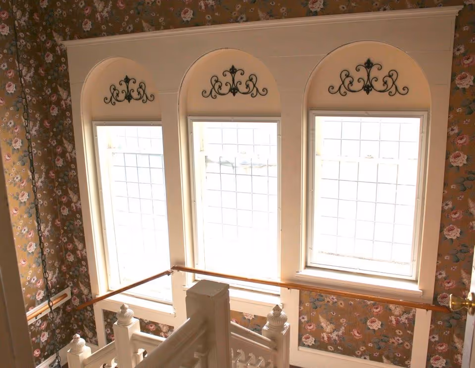 Stairwell landing with three tall arched windows, decorative iron scrollwork, floral wallpaper, and a wooden banister.