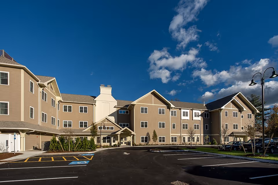 Exterior view of a large, three-story senior living facility building under a blue sky with some clouds. The building has beige siding with white trim and multiple windows. There is a parking lot with marked spaces, including handicapped spots, and some small trees and shrubs near the entrance.