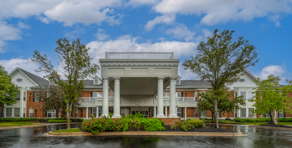 Front exterior view of a senior living facility named Waltonwood Twelve Oaks, featuring a large covered entrance with white columns, red brick walls, white trim, and several windows. The driveway is wet from rain, and there are trees and shrubs around the entrance under a partly cloudy blue sky.