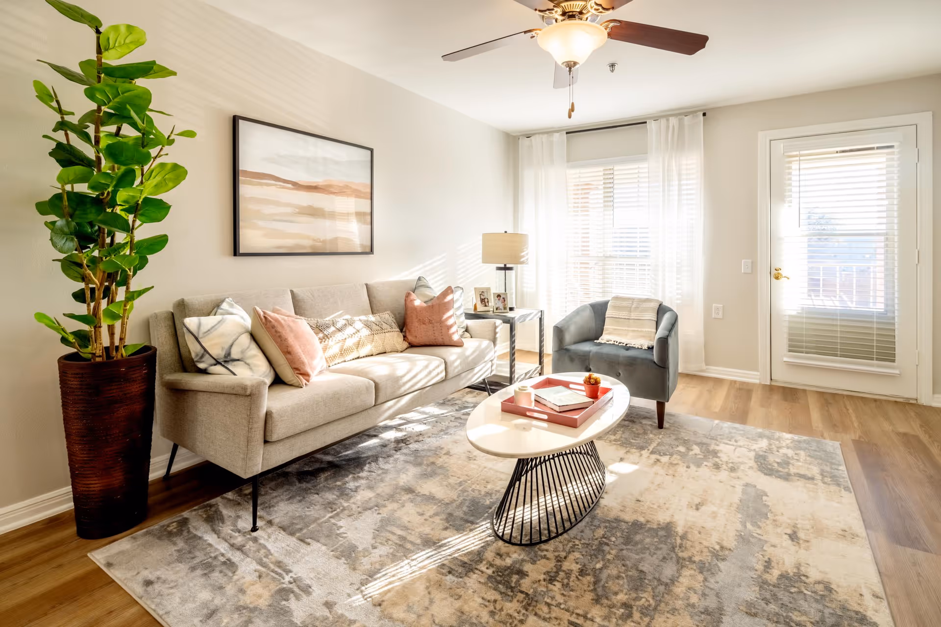 A bright living room with a beige sofa adorned with patterned and solid pillows, a gray armchair with a throw blanket, a modern oval coffee table with a red tray and decorative items, a tall potted plant, a floor lamp, and large windows with white curtains allowing natural light to fill the space.